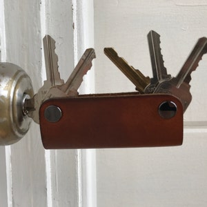 May include: A brown leather key holder with silver metal accents and multiple keys attached. The key holder is attached to a door knob.