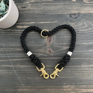 Puede incluir: Correa doble para perro negra hecha de cuerda gruesa con dos clips de metal dorado y un anillo dorado. La correa tiene forma de corazón.