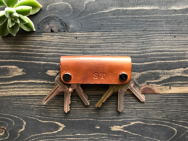 May include: A brown leather key holder with a silver snap closure and the initials "ST" embossed on the front. The key holder is holding a set of keys.