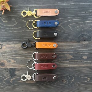May include: Seven leather keychains with metal rings and clasps. Each keychain has a different color and is personalized with a name: MOM, DAD, JR, NR, CWF, TGM, and MB.