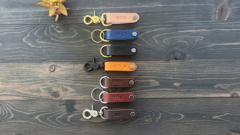 May include: Seven leather keychains with metal rings and clasps. Each keychain has a different color and is personalized with a name: MOM, DAD, JR, NR, CWF, TGM, and MB.