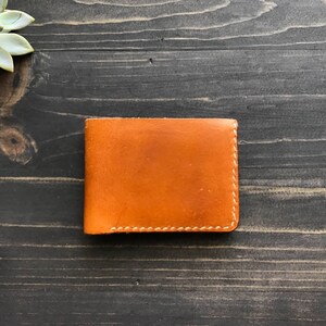 May include: A brown leather wallet with white stitching. The wallet is closed and lying flat on a dark wood surface.