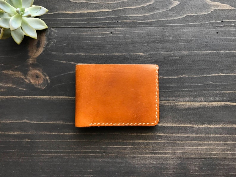 May include: A brown leather wallet with white stitching. The wallet is closed and lying flat on a dark wood surface.