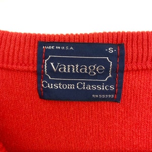 May include: Close-up of a red knit fabric with a navy blue label. The label reads "Vantage Custom Classics" with "Made in U.S.A." above and "RN 55393" below. The label is stitched with red thread.