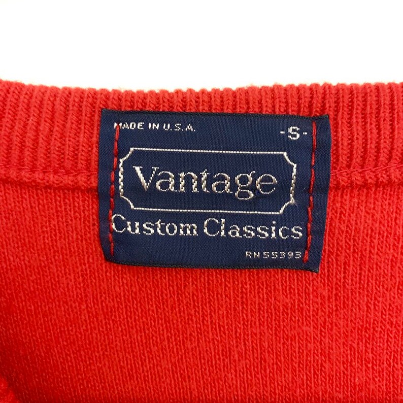 May include: Close-up of a red knit fabric with a navy blue label. The label reads "Vantage Custom Classics" with "Made in U.S.A." above and "RN 55393" below. The label is stitched with red thread.
