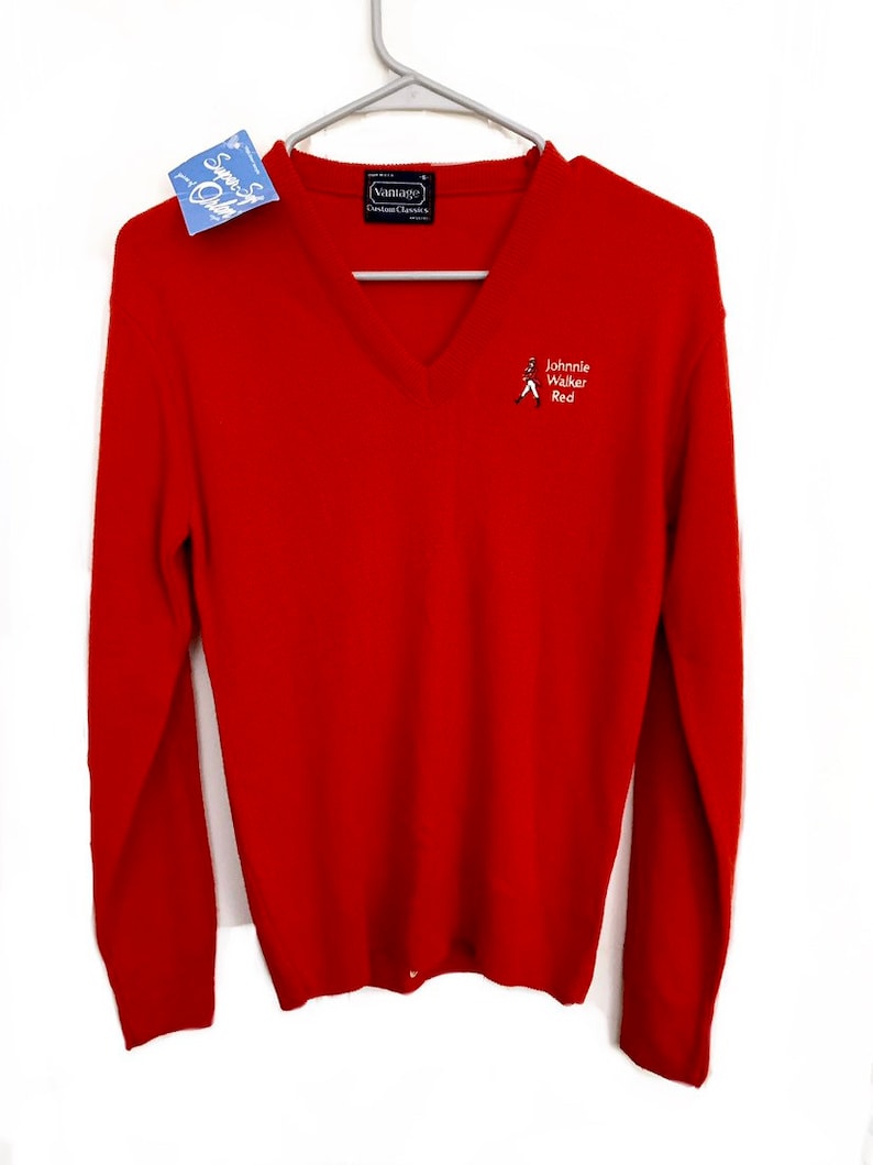 May include: A red V-neck sweater with long sleeves, hanging on a hanger. The sweater has the text "Johnnie Walker Red" embroidered on the chest, along with a logo. A tag is attached to the sweater.