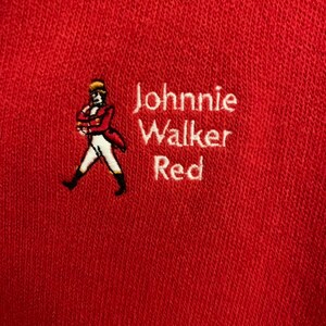 May include: Red fabric with embroidered text and a logo. The text reads "Johnnie Walker Red" in white. The logo is a man in a red coat and white pants. The fabric is a solid red color.