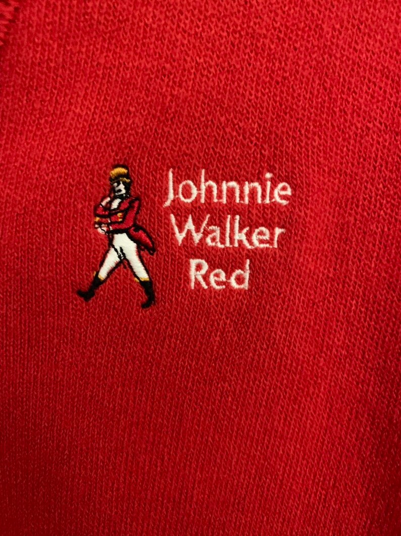May include: Red fabric with embroidered text and a logo. The text reads "Johnnie Walker Red" in white. The logo is a man in a red coat and white pants. The fabric is a solid red color.