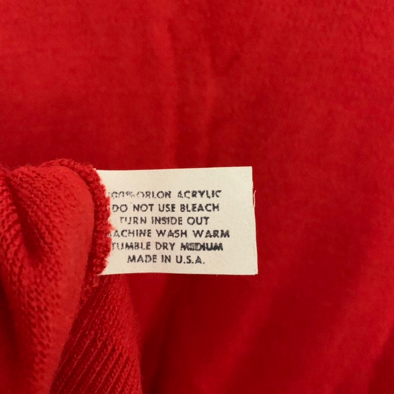 May include: Close-up of a red garment with a white tag attached. The tag contains care instructions and the text "100% ORLON ACRYLIC, DO NOT USE BLEACH, TURN INSIDE OUT, MACHINE WASH WARM, TUMBLE DRY MEDIUM, MADE IN U.S.A."