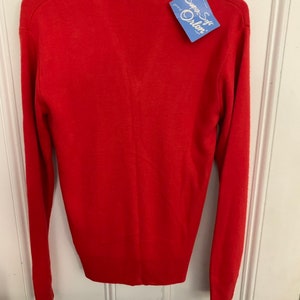 May include: A bright red sweater with long sleeves, hanging on a hanger. The sweater has a crew neck and a tag that reads "Super Soft Orlon". The garment is made of a soft material.