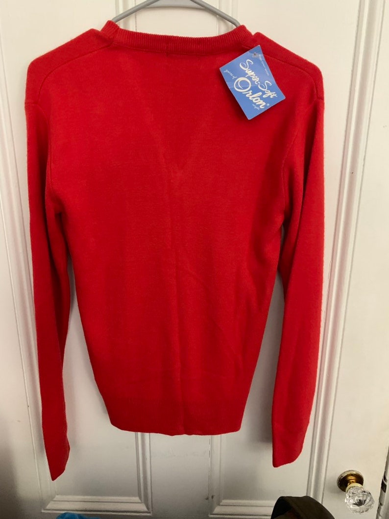 May include: A bright red sweater with long sleeves, hanging on a hanger. The sweater has a crew neck and a tag that reads "Super Soft Orlon". The garment is made of a soft material.