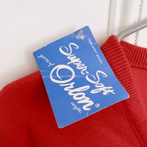 May include: A rust-colored sweater with a blue tag that reads "Super-Soft Orlon acrylic." The tag also states "100% Orion acrylic" and "yarns of". The sweater is hanging on a metal hanger against a white background.