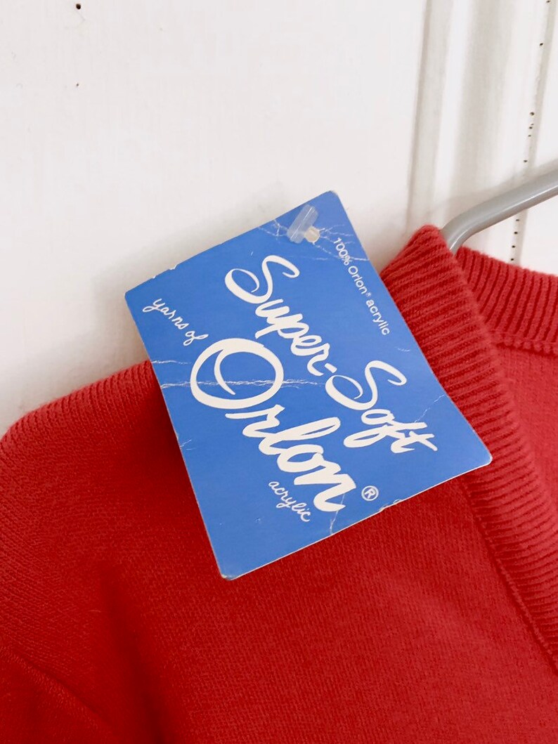 May include: A rust-colored sweater with a blue tag that reads "Super-Soft Orlon acrylic." The tag also states "100% Orion acrylic" and "yarns of". The sweater is hanging on a metal hanger against a white background.