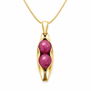 May include: A gold-toned pendant necklace featuring a stylised pea pod shape holding two round, polished magenta-coloured beads. The pendant is suspended from a thin, matching gold-toned snake chain, creating a cohesive and polished jewellery piece.