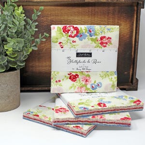 May include: A stack of fabric squares featuring a floral design in shades of pink, red, blue, and green on a white background. The fabric squares are labelled "Hollyhocks & Roses" by Bunny Hill Designs.