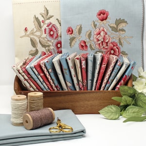 May include: A wooden tray filled with folded fabric squares featuring floral patterns in red, blue, and cream. Three wooden spools of thread in white, tan, and brown, a pair of gold scissors, and a stack of solid light blue fabric are included.