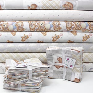 May include: A collection of folded and rolled cotton fabric featuring repeating patterns of brown teddy bears, pastel balloons, stars, clouds, and moons in shades of beige, light blue, and soft pink. The fabric stacks are tied with white ribbons.