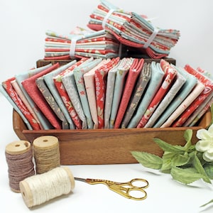 May include: Fabric bundles in red, blue, and grey, displayed in a wooden container. Some bundles are tied with white ribbon. Spools of thread and gold scissors are also present, indicating a sewing or quilting theme, ideal for craft projects.
