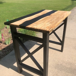 May include: A handcrafted wooden table with a light-colored top and dark, black epoxy resin inlays. The table has dark brown legs with an X-shaped support structure. The table is approximately 40 inches long and 20 inches wide.