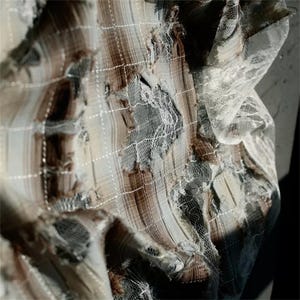 May include: Close-up of a distressed textile with a layered design. The fabric features brown, grey, and cream tones, with a grid-like pattern of white stitching. Sections of the fabric are torn, revealing layers of lace and other materials.