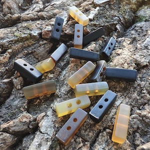 May include: A collection of rectangular toggle buttons featuring two holes for attachment. The buttons are rendered in a variety of colors, including solid black, dark brown, and translucent amber, each with a smooth, polished finish and rounded edges.