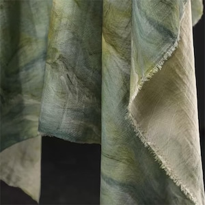 May include: Close-up of flowing fabric in shades of green and white. The material has a textured appearance with a subtle, watercolor-like pattern. The edges of the fabric are frayed, adding a rustic touch.