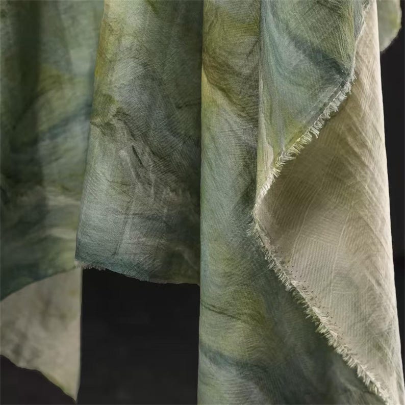 May include: Close-up of flowing fabric in shades of green and white. The material has a textured appearance with a subtle, watercolor-like pattern. The edges of the fabric are frayed, adding a rustic touch.