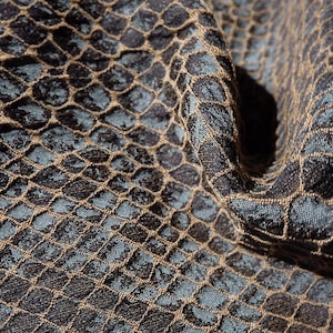 Puede incluir: Primer plano de una tela texturizada con un patrón geométrico repetitivo. La tela presenta un fondo verde azulado oscuro con una superposición marrón en forma de cuadrícula, creando un efecto dimensional. La tela está doblada, mostrando profundidad y textura.