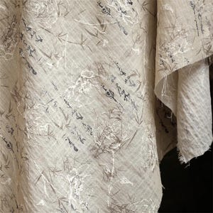 Op de afbeelding: Een beige stof met een geprint patroon van donkerbruine bamboestengels en zwart kalligrafisch schrift. Het textiel toont witte, verhoogde, draadachtige bloemcontouren die over de print zijn aangebracht, met gerafelde randen aan de zijkant.