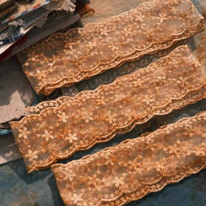 Puede incluir: Primer plano de varias tiras de encaje delicado, de color marrón claro. El encaje presenta un patrón floral y un borde festoneado. Las tiras están dispuestas sobre una superficie azul y beige, lo que sugiere un proyecto de artesanía o costura.