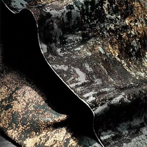 May include: Close-up of a textured fabric with a metallic sheen. The fabric features a dark base color with silver and gold accents, creating a layered, abstract pattern. The material is folded, revealing a deep black interior.