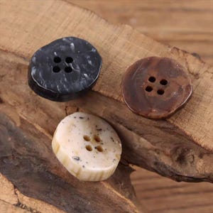 May include: Three decorative buttons in a close-up shot. The buttons are in shades of black, brown, and cream. Each button has four holes for sewing. The buttons are made of a smooth, possibly polished material.