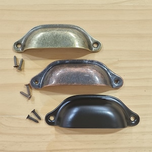 Puede incluir: Tres tiradores de armario tipo taza en acabados de latón antiguo, cobre y negro. Cada tirador tiene dos orificios para tornillos y va acompañado de tornillos a juego. Los tiradores están dispuestos sobre una superficie de madera clara.