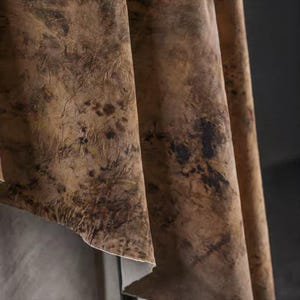 May include: Close-up of a textured fabric with a mottled appearance. The material is primarily brown with darker brown and black splotches, creating a marbled effect. The fabric is draped, showing folds and edges.