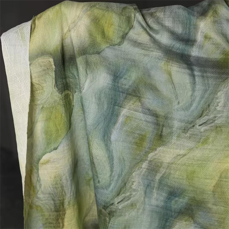 May include: A draped textile with a watercolor-like design. The fabric features a blend of green, blue, and white hues, creating a soft, flowing appearance. The texture appears delicate, with subtle variations in color and tone.