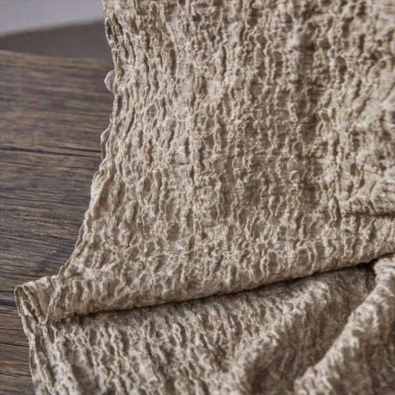 May include: Close-up of a beige, textured fabric with a crinkled appearance. The fabric has a woven texture and is folded over, revealing the edge of the material. The color is a neutral tone, suitable for various home decor applications.