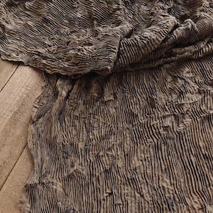 May include: A textured, brown fabric with vertical, ribbed lines. The fabric is draped over a wooden surface, creating folds and shadows. The material appears to be soft and flowing, with a slightly wrinkled appearance.
