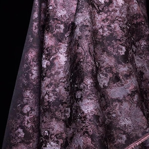 May include: Close-up of a draped fabric with a marbled pattern in shades of purple, black, and white. The fabric has a soft, flowing texture, with visible folds and subtle variations in color and tone. The overall effect is a blend of elegance and artistic design.