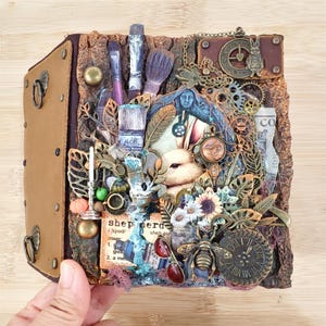 May include: A mixed media art piece featuring a rabbit, clock, and bee. The artwork incorporates paintbrushes, gears, and botanical elements. The piece is encased in a brown leather-bound book with metal hardware.
