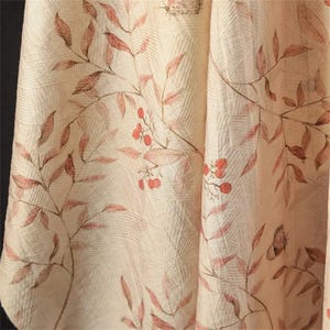 Op de afbeelding: Close-up van een cr&egrave;mekleurige stof met een botanische print. Het ontwerp toont takken met roodbruine bladeren en kleine rode bessen. Subtiele vlinderillustraties zijn ook zichtbaar, wat een natuurlijke, elegante esthetiek cre&euml;ert.