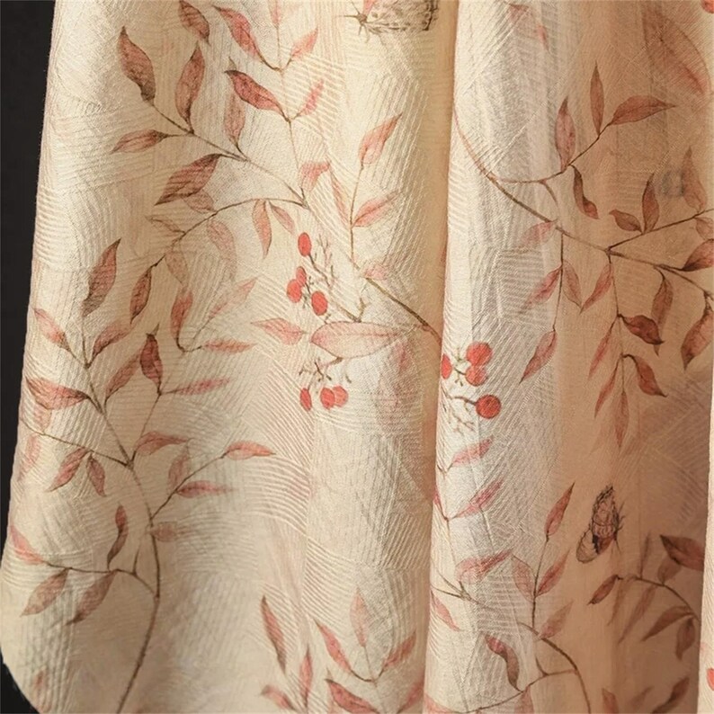 Op de afbeelding: Close-up van een cr&egrave;mekleurige stof met een botanische print. Het ontwerp toont takken met roodbruine bladeren en kleine rode bessen. Subtiele vlinderillustraties zijn ook zichtbaar, wat een natuurlijke, elegante esthetiek cre&euml;ert.