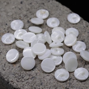 Puede incluir: Un conjunto de botones blancos y redondos con un brillo nacarado. Los botones están dispersos sobre una superficie rugosa y gris. Cada botón tiene una calidad sutil e iridiscente, con un diseño de dos agujeros.