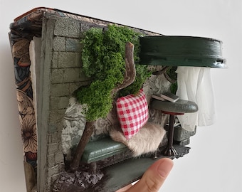 Könnte beinhalten: Miniatur-Diorama einer gemütlichen Außenszene, eingebettet in die Seiten eines Buches. Mit einer grünen Bank, einem rot-weiß karierten Kissen, einem kleinen Baum und einem kleinen Tisch mit Büchern. Der Buchdeckel hat ein Blumenmuster.