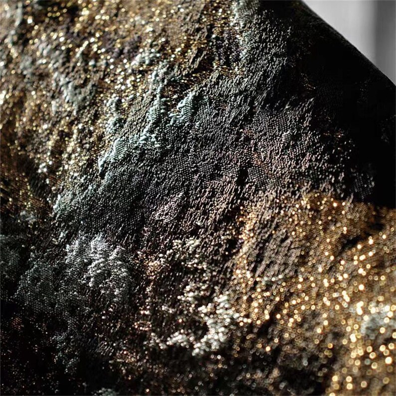 May include: Close-up of a textured fabric with a shimmering, metallic appearance. The fabric features a blend of black, gold, and silver tones, creating a luxurious and detailed surface. The light catches the texture, highlighting the intricate weave.