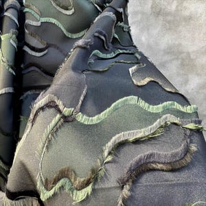 May include: Close-up of a dark fabric with wavy, fringed edges in shades of green, gray, and black. The fabric has a textured appearance, with the fringing adding a tactile element to the design. The overall aesthetic is modern and artistic.