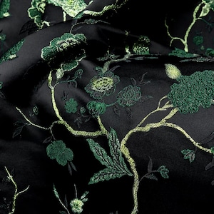 May include: A black fabric featuring an intricate embroidered pattern of flowers and branches in shades of dark green, lime green, and metallic gold. The embroidery has a textured, raised appearance against the smooth, dark satin-like material.