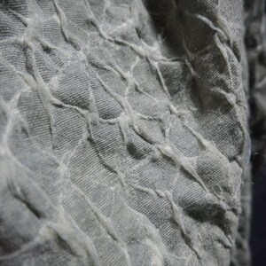 May include: Close-up of a textured fabric in shades of gray. The fabric has a quilted appearance with raised, irregular lines creating a grid-like pattern. The texture appears soft and slightly wrinkled, with a subtle sheen.