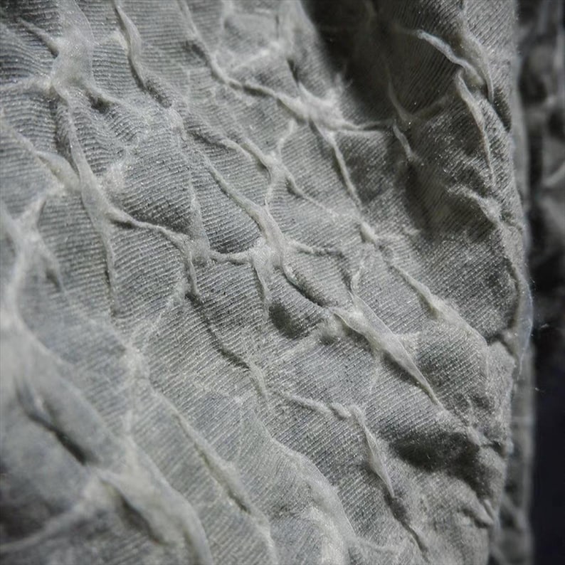 May include: Close-up of a textured fabric in shades of gray. The fabric has a quilted appearance with raised, irregular lines creating a grid-like pattern. The texture appears soft and slightly wrinkled, with a subtle sheen.
