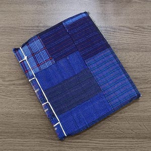 Peut inclure: Un journal fait à la main avec une couverture patchwork dans différentes nuances de bleu. La couverture est faite de carrés de tissu cousus ensemble, avec du fil blanc reliant les pages. Le journal est rectangulaire et repose sur une surface en bois.