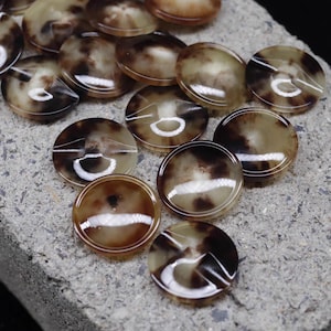 May include: A collection of round, brown and tan buttons with a marbled appearance. Each button has a white, circular groove around the edge. The buttons are arranged on a gray, textured surface. These buttons are suitable for sewing and craft projects.